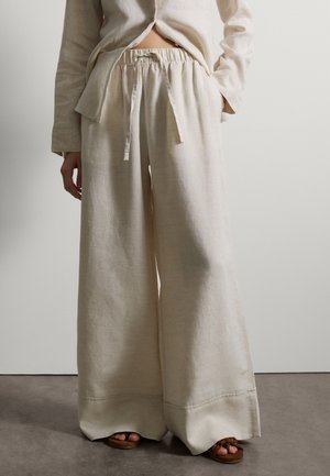 Persona che indossa pantaloni larghi in lino beige con vita con coulisse e camicia abbinata, in piedi su un pavimento grigio contro una parete bianca.