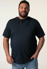 Tmavě modrá košile s krátkým rukávem a šněrovacím dekoltem. Měkká, texturovaná tkanina s uvolněným střihem a zakulaceným spodním okrajem. Nosená s světle modrými džíny.