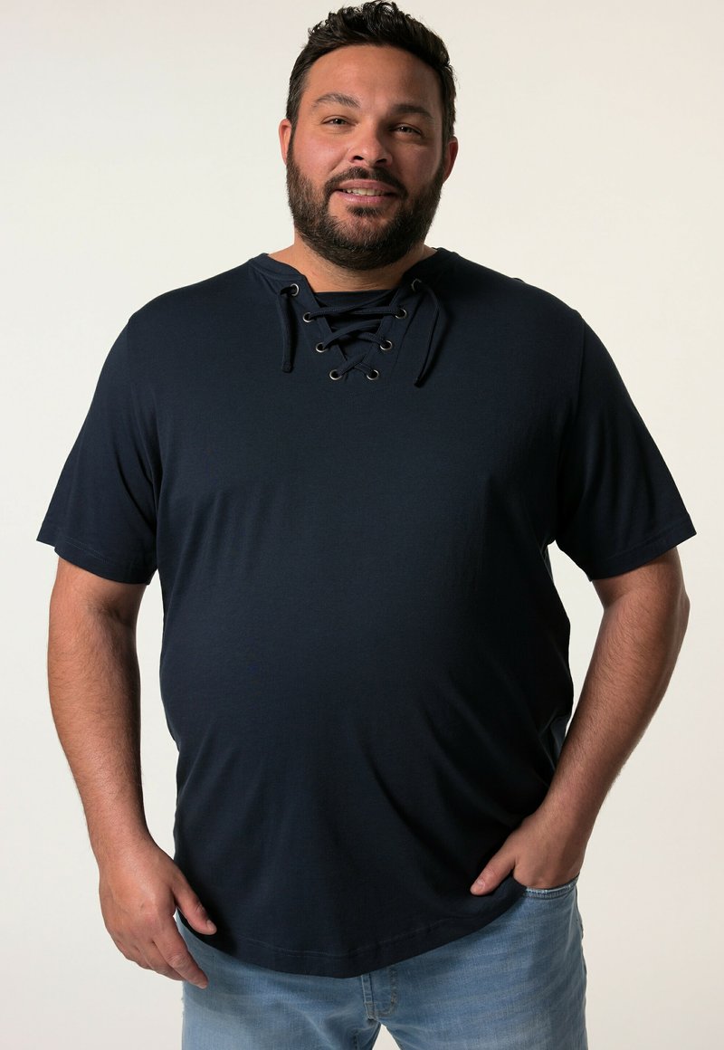 Tmavě modrá košile s krátkým rukávem a šněrovacím dekoltem. Měkká, texturovaná tkanina s uvolněným střihem a zakulaceným spodním okrajem. Nosená s světle modrými džíny.