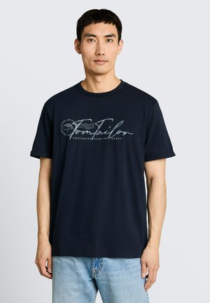 Młody mężczyzna w granatowym T-shircie z napisem „Tom Tailor” i jasnoniebieskich dżinsach, stojący na jednolitym jasnoszarym tle.