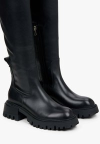 Knee-high black leather boots with a smooth texture, side zipper, and chunky rubber sole featuring a ridged pattern for traction.
