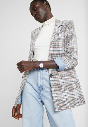 Kobieta w białym golfie, niebieskiej marynarce w kratę z podwiniętymi rękawami, jasnoniebieskich dżinsach z wysokim stanem i zegarku na rękę z niebieskim paskiem.