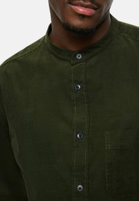 Olivgrünes Cordhemd mit Stehkragen, knöpfbarem Frontverschluss, schwarzen Knöpfen und einer Brusttasche. Glatte Textur mit vertikalen Linien.