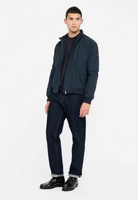 Marineblaue Bomberjacke mit Reißverschluss, gerippten Bündchen und Seitentaschen, kombiniert mit dunklen Denim-Jeans und schwarzen Schuhen.