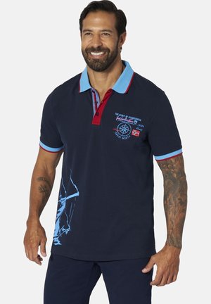 Man smiling wearing navy blue polo with light blue and red collar and printed compass design on chest and sleeve tattoos visible.