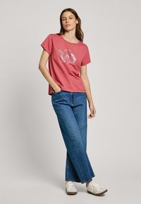 T-shirt rouge à manches courtes avec un design graphique argenté sur le devant, associé à un jean en denim bleu ample et des baskets blanches.