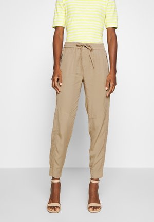 Pantalons beige à cordon de serrage en tissu léger, avec des poches latérales et des chevilles fuselées. Portés avec un haut à rayures jaunes et des talons.