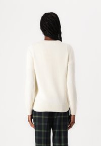Maglione bianco a coste con maniche lunghe, scollo rotondo e texture intrecciata. Il modello indossa pantaloni a quadri con motivi verdi e neri.