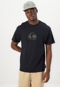 Black short-sleeve t-shirt with a tonal Quiksilver logo. Simple, crew neck design. Worn with light beige shorts and a gray cap.