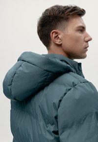 Giacca piumino in verde scuro con cappuccio morbido, texture liscia e design trapuntato. Caratterizzata da un alto colletto e elementi stilistici minimalisti.