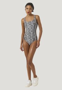 Kvinna stående med benen korsade iförd en svart och vit ormtryckt baddräkt och vita sneakers mot en enkel bakgrund.