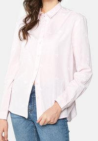 Ljusrosa skjorta med knappar, tillverkad av bomull med vita broderade palmträdsmönster; har långa ärmar och klassisk krage.