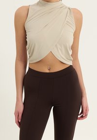 Beżowy top bez rękawów z wysokim kołnierzem, ozdobiony drapowaną tkaniną i skręconym przodem, połączony z brązowymi legginsami z wysokim stanem.