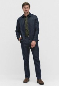 Granatowa kurtka dżinsowa i pasujące spodnie z zieloną koszulą z kołnierzykiem pod spodem. Zawiera kieszeń na piersi, prosty krój i brązowe buty.