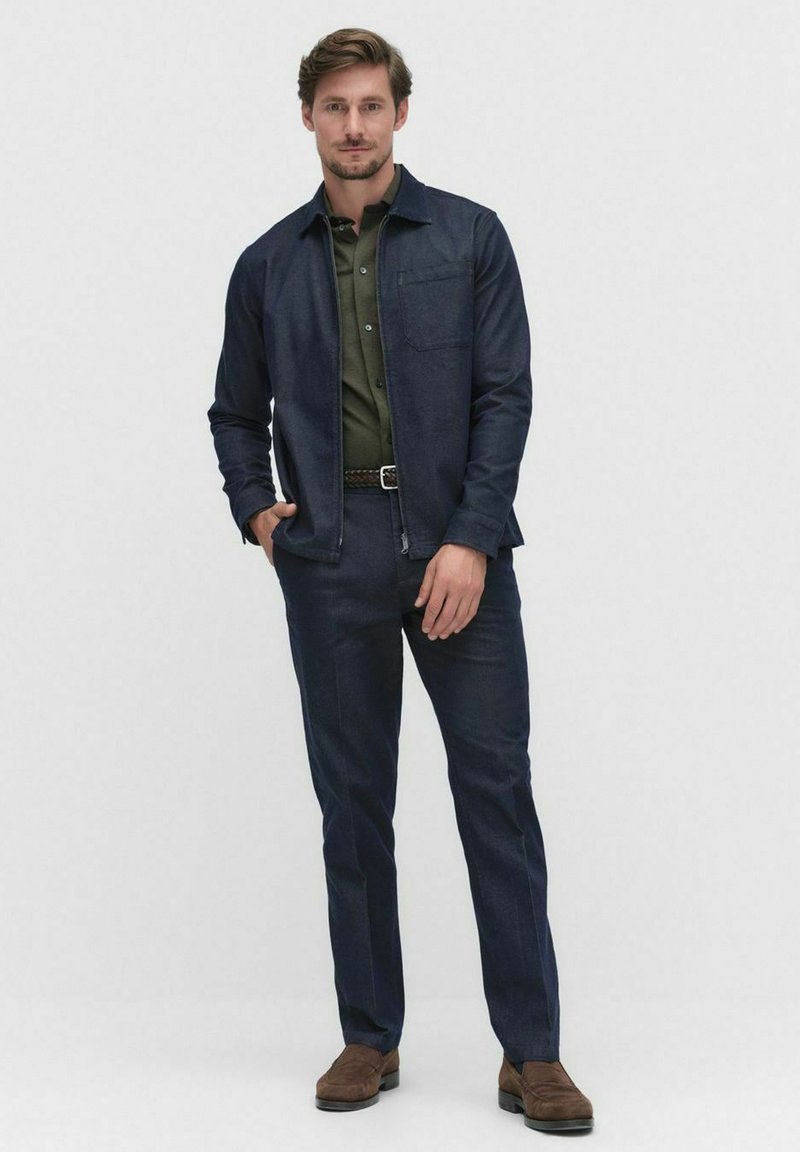 Granatowa kurtka dżinsowa i pasujące spodnie z zieloną koszulą z kołnierzykiem pod spodem. Zawiera kieszeń na piersi, prosty krój i brązowe buty.