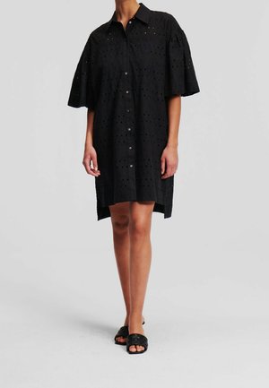 Femme portant une robe noire ample boutonnée avec des manches larges et un motif perforé, associée à des sandales slides tissées noires à bout ouvert.