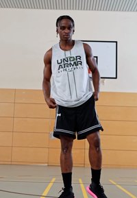 Atleta con una camiseta sin mangas gris de Under Armour con logo verde, pantalones cortos negros con detalles en blanco y zapatillas deportivas negras.