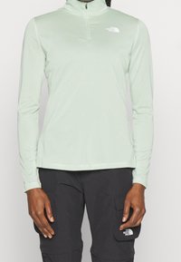 Jasnozielona, długosleeve'owa koszula z zamkiem na szyi, wykonana z gładkiego materiału. Ma małe białe logo na lewej stronie klatki piersiowej.