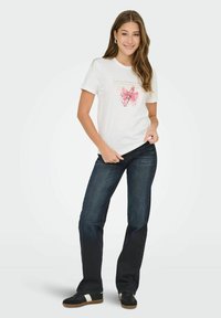 Vrouw glimlachend in een wit t-shirt met een roze strik graphic, donkere spijkerbroek en zwarte sneakers met witte strepen, staand op een witte achtergrond.