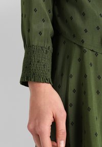 Manche de robe verte avec motif à losanges noirs, dotée d'un poignet froncé, tissu lisse et légère brillance. Main visible sous la manche.