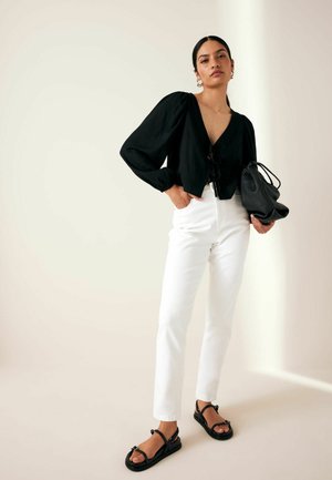 Blouse noire courte avec des manches bouffantes, décolleté en V et détail à nouer ; associé à un jean blanc droit et des sandales noires. Sac noir tenu à la main.