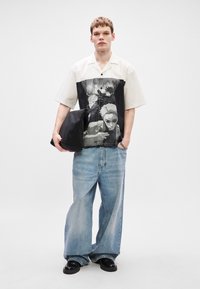 Το κοντομάνικο πουκάμισο διαθέτει μια μονοχρωμία γραφική εκτύπωση στο μπροστινό μέρος, συνδυασμένο με φαρδιές ανοιχτό μπλε τζιν και μαύρα παπούτσια. Μαύρη τσάντα στο χέρι.