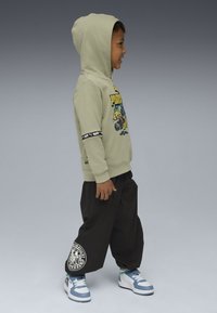 Ragazzo in felpa grafica verde chiaro, pantaloni neri larghi con toppe e sneakers blu-bianche, in piedi di lato, sorridendo su sfondo grigio.
