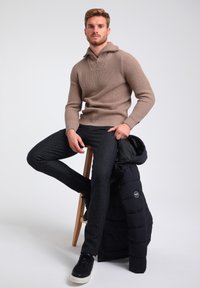 Beige gerippter Pullover mit umgeschlagenem Kragen, dunklen Textilhosen und schwarzer Steppjacke, die über einem Holzstuhl hängt. Schwarze Sneaker.