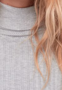 Cheveux blonds ondulés tombant sur un pull à col roulé côtelé gris clair, révélant la zone du cou et des épaules.
