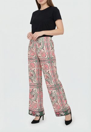 Mujer con una blusa negra de manga corta, pantalones de pierna ancha de cintura alta con un patrón geométrico floral en rosa, verde y beige, y tacones negros.