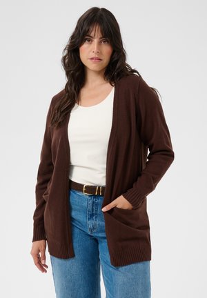 Vrouw met lang donker haar draagt een bruine cardigan, een wit topje, een blauwe spijkerbroek en een zwarte riem, staand met één hand in haar zak.