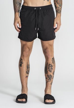 Zwarte zwemshorts met elastische tailleband en trekkoord. Licht, soepel materiaal. Draag met zwarte gestructureerde slippers. Zichtbare been tattoo's.
