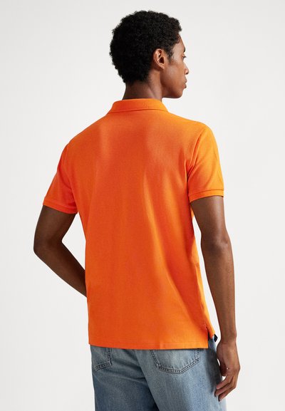 Polo orange avec un tissu texturé, manches courtes et col. Le dos présente un ourlet légèrement plus long et des fentes latérales pour plus de confort.