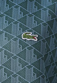 Grön stickad pikétröja med ett geometriskt blått mönster. Den framträdande Lacoste-krokodillogotypen är broderad i grönt och rött på bröstet.