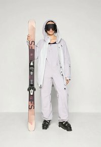 Σαπούνι σκι που περιλαμβάνει ελαφρύ γκρι αδιάβροχο μπουφάν, ταιριαστές φόρμες, μαύρες μπότες σκι και ένα ζευγάρι σκι με δέστρες.