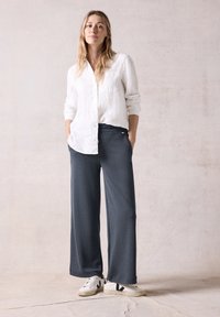 Weiße langärmelige Leinenbluse, graue weit geschnittene Hose und weiße Sneakers mit schwarzen Akzenten. Das Modell steht mit den Händen in den Taschen vor einem neutralen Hintergrund.