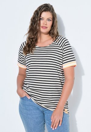 Frau mit langem, welligem Haar trägt ein kurzärmeliges, schwarz-weiß gestreiftes Shirt mit pfirsichfarbenen Akzenten und blaue Jeans, sie steht vor einer weißen Wand.