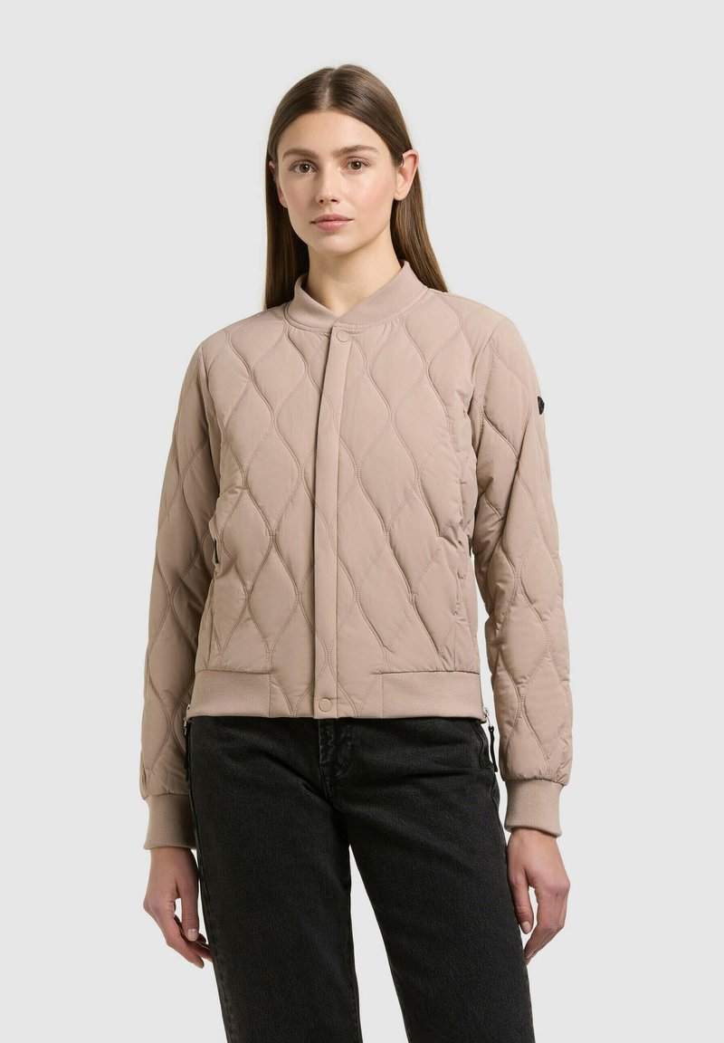 Femme aux cheveux bruns raides portant une veste bomber matelassée beige et un pantalon noir, debout devant un fond clair uni.