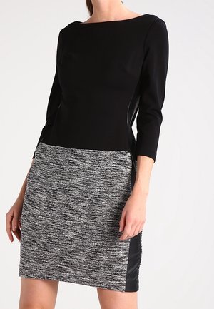 Femme portant une robe texturée noire et grise à manches trois-quarts, debout devant un fond blanc uni.