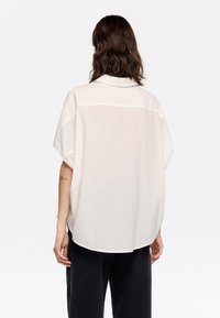 White textured shirt with a relaxed fit, short sleeves, and a back yoke detail, paired with black wide-leg pants.