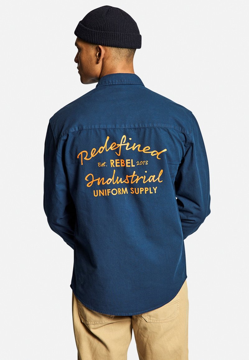 Camicia a maniche lunghe di un blu navy con testo arancione sulla schiena, che riporta "Redefined," "REBEL 2013," e "Industrial UNIFORM SUPPLY." Miscela di cotone, texture morbida.