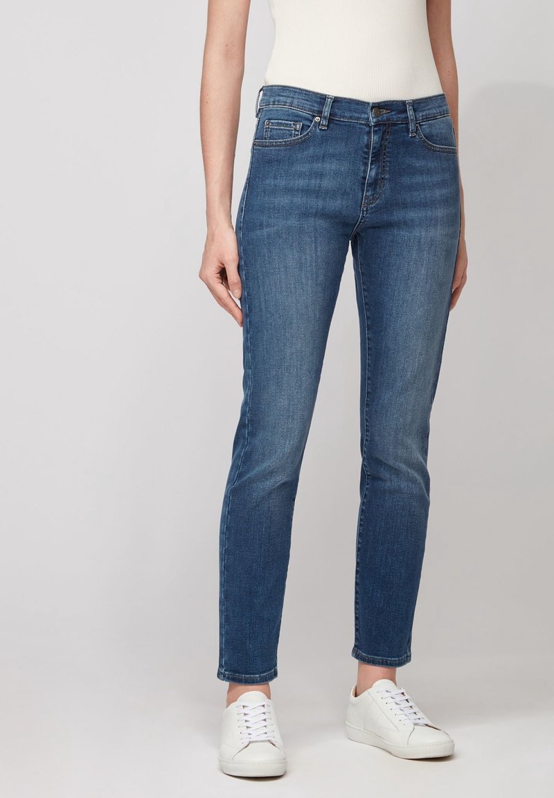 Femme portant un jean bleu slim taille moyenne, des baskets blanches et un top blanc sans manches, debout devant un fond clair uni.