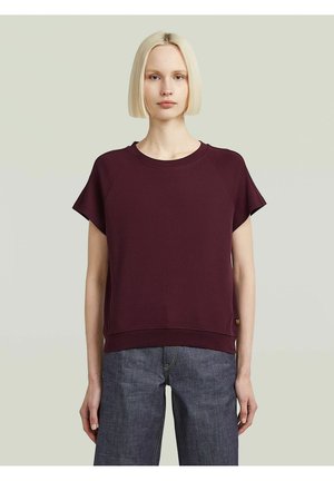 Jeune femme aux cheveux blonds courts, portant un haut à manches courtes bordeaux foncé et un jean bleu, sur un fond clair uni.