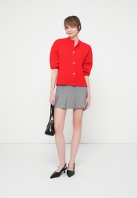 Femeie îmbrăcată cu un pulover roșu cu nasturi, fustă mini plisată gri, pantofi cu toc negri și ținând o geantă neagră, stând în fața unui fundal alb.