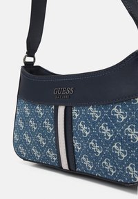 Blaue Jeans und marineblaue Schultertasche mit weißen gemusterten Logos, vertikalen weißen und marineblauen Streifen sowie silberner „GUESS EST 1981“-Prägung.