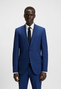 Blauw op maat gemaakt pak met een slim fit, voorzien van een enkelgeknoopt jasje, notched revers en twee voorzakken. Draagt over een geblokte witte blouse.