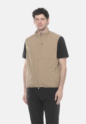 Uomo con capelli corti e scuri che indossa un gilet beige con zip sopra una camicia nera a maniche corte e pantaloni neri, in piedi davanti a uno sfondo chiaro.