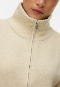 Maglione cream con zip, dotato di un alto colletto a coste, con una zip argentata e un tessuto morbido e strutturato. Design semplice e pulito.