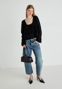 Giovane donna che indossa una blusa nera con pizzo, jeans blu, tacchi a punta neri e orecchini a cerchio, mentre tiene in mano una piccola borsa nera.