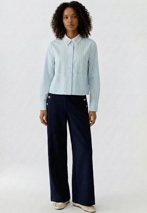 Frau steht und trägt ein hellblau gestreiftes Hemd mit weißen Kragen und eine marineblaue Hose mit weitem Bein und goldenen Knöpfen.